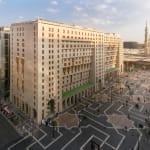 Außenansicht des Sofitel Shahd Al Madinah Hotels bei Tageslicht, mit einem großen Platz und Menschen, die sich bewegen.