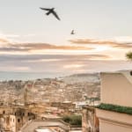A cityscape view from a rooftop terrace in Fez during sunset, with birds flying across the sky and mountains in the background.