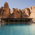 Ein Swimmingpool mit einem modernen Gebäude im Hintergrund, umgeben von felsigem Wüstenland während des Tages.