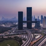 Panoramablick auf eine Stadt bei Dämmerung mit zwei hohen dunklen Wolkenkratzern, die durch eine Himmelbrücke verbunden sind, umgeben von Autobahnen, Grünflächen und zahlreichen anderen Gebäuden.