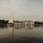 A large white riverboat with multiple decks sails on a calm river during the day, accompanied by a smaller traditional sailboat with a tall white sail, both reflected in the water.