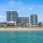 Ein modernes Strandhotel mit mehreren Stockwerken und Glasfassade, gelegen an einem tropischen Strand mit Palmen.