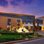 Außenansicht des Hotels St. Regis Riyadh mit beleuchteter Fontäne und gestalteten Gärten bei Dämmerung.