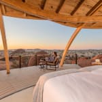 Eine Person sitzt auf einem Stuhl auf einer hölzernen Terrasse und genießt den Sonnenuntergang über einer Wüstenlandschaft mit Felsen und spärlicher Vegetation.