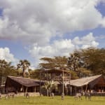 A main lodge area at Chem Chem Lodge in Tanzania with zebras grazing in the foreground under a partly cloudy sky.