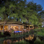 A luxurious guest suite located outdoors beneath large trees, featuring a private deck with a hot tub, illuminated during twilight in a natural setting.