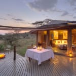 Luxurious outdoor dining area on a wooden deck at sunset, overlooking a savannah landscape, with a tent structure illuminated from within.