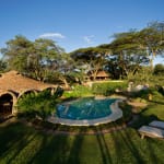 A scenic view of a garden with a round swimming pool, lush green trees, and thatched-roof structures under a clear blue sky.