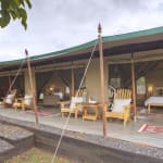 Ein großes Familienzelt mit einer geräumigen offenen Front, ausgestattet mit Holzmöbeln wie Stühlen, Sofas und Betten im Inneren, in einer natürlichen Außenumgebung.