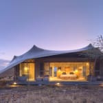 Luxurious tent with warm interior lighting set in a dry, open landscape at dusk, framed by trees.
