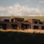 A series of modern, dark wood-paneled buildings set against a backdrop of grassy hills under a partly cloudy sky during sunset.