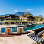 A modern outdoor swimming pool area with multiple lounge chairs and umbrellas, set against a backdrop of wooden cabins and a mountainous landscape under a clear blue sky.