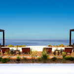 A luxurious outdoor terrace with two canopy daybeds and several wooden lounge chairs facing a calm ocean under a clear blue sky.