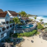 Coastal resort with triangular-roofed buildings, central round swimming pool, palm trees, and sandy beach adjacent to the ocean under a clear blue sky.