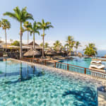 A tropical resort pool area with two adjacent infinity pools, palm trees, thatched umbrellas, and lounge chairs overlooking the ocean under a clear blue sky.