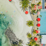 Luftaufnahme eines Resorts mit Sandstrand, klarem türkisfarbenem Wasser, Palmen und einem Schwimmbad mit roten Sonnenschirmen.