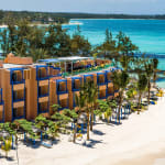 Orange-colored beachfront hotel with blue window frames situated along a white sandy beach with palm trees and turquoise ocean water in the background.