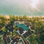 Aerial view of a tropical resort with a large curvy swimming pool surrounded by umbrella-shaded loungers, buildings with dark roofs, dense greenery, and a sandy beach adjacent to calm sea water reflecting sunlight.