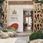 Eingang zu Myconian Naia Luxury Suites umgeben von Bäumen und Felsen, mit einer modernen Dekorationswand und sichtbarem Innenraum durch die Tür.