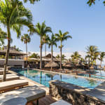Outdoor pool area of a tropical hotel or spa with lounge chairs, palm trees, and clear blue sky by the sea.