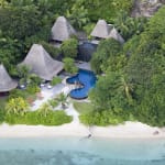 Aerial view of a tropical resort with thatched-roof villas, a swimming pool, and a white sandy beach surrounded by lush green trees.