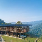 A modern glass building with a sloped roof and large windows overlooking a lush green valley and hills under a clear blue sky.
