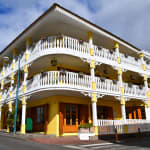 A large yellow two-story building with ornate white railings and balconies on both floors, decorated with hanging flower pots, situated at a street corner under a partly cloudy sky.