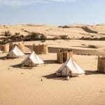 A desert scene with several white tents and small wooden structures on sand dunes under a blue sky.