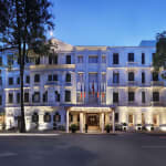 The exterior of the Sofitel Legend Metropole Hanoi hotel illuminated at dusk, with trees and a street in front.