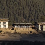Three traditional Bhutanese buildings with intricate woodwork sit in front of a forested mountain.