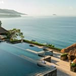 Ein mehrstufiger Infinity-Pool mit Blick auf den Ozean, mit Strohdachhütten in der Nähe und üppigem Grün im Hintergrund.