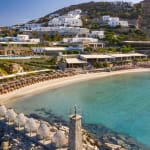 Ein malerischer Blick auf den Strand und den Resortbereich in Santa Marina auf Mykonos, Griechenland, mit weißen Gebäuden an einem Hang und Liegestühlen am Ufer.