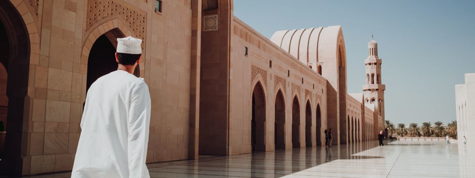 Ein Mann in weißer traditioneller Kleidung spaziert durch den geräumigen Hof einer großen Moschee, mit aufwändiger Architektur und einem hohen Minarett im Hintergrund.