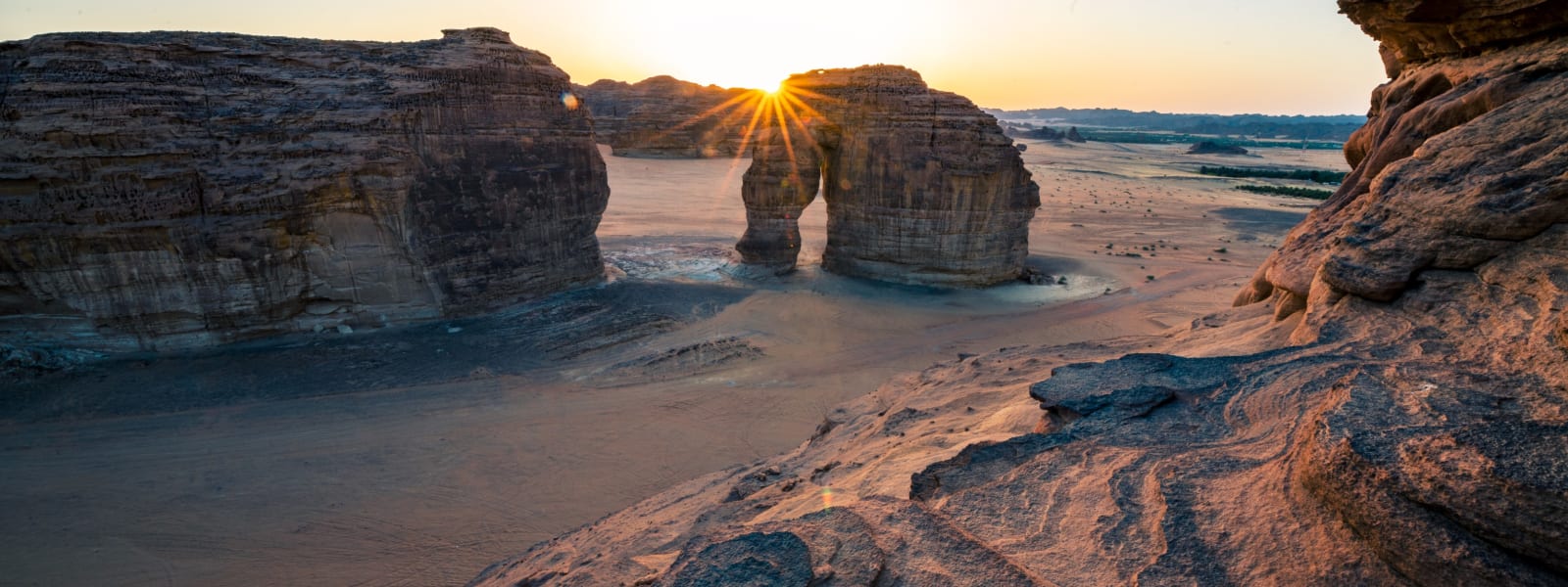 Sunset over unique stone formations in AlUla, Saudi Arabia, with sun rays peeking through a natural arch.