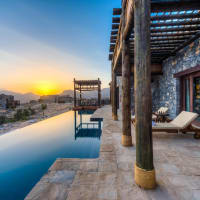 A luxurious stone villa with a private infinity pool overlooking a sunset landscape with mountains.
