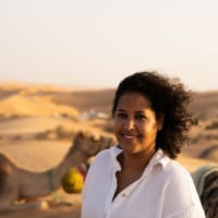 Eine Frau mit lockigem Haar und weißem Hemd lächelt in einer Wüstenlandschaft mit Kamelen und Sanddünen im Hintergrund bei Sonnenuntergang.