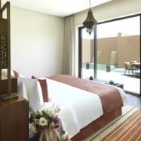 Ein geräumiges Hauptschlafzimmer mit einem großen Bett, dekorativen Kissen und einer Vase mit Blumen, das zu einer Terrasse mit Poolmöbeln und Gartenblick führt.
