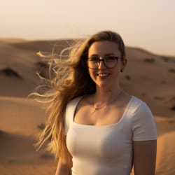 Junge Frau mit langen blonden Haaren und Brille, die in einer Wüstenlandschaft bei Sonnenuntergang lächelt.