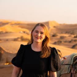 A woman with blonde hair and a black dress smiling in a desert landscape with sand dunes and camels in the background during sunset.
