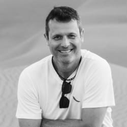 Smiling man with short hair wearing a white t-shirt and sunglasses hanging from the collar, posing outdoors with a blurred sandy background.