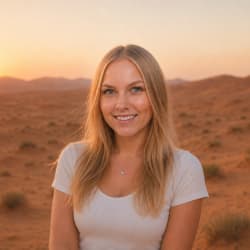 Lächende junge Frau mit langen blonden Haaren, die ein weißes Kurzarmshirt trägt und in einer Wüstenlandschaft bei Sonnenuntergang steht.