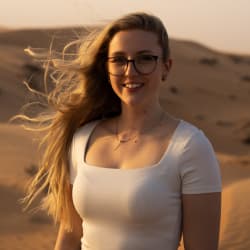 A smiling woman with long blonde hair and glasses wearing a white top standing in a desert during golden hour.