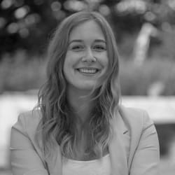 A young woman with long wavy hair smiling outdoors with a blurred natural background.