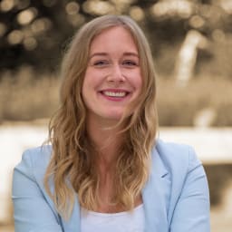 Eine junge Frau mit langen gewellten blondem Haar, die im Freien lächelnd trägt, in einem hellblauen Blazer.