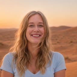 A smiling woman with long blonde hair wearing a light blue shirt standing in the desert during sunset with sand dunes in the background.