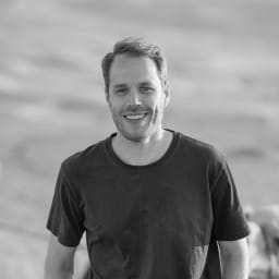A smiling man with short hair and a beard, wearing a dark t-shirt, standing outdoors.