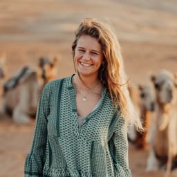 A smiling woman with wavy hair standing outdoors with camels in the background.