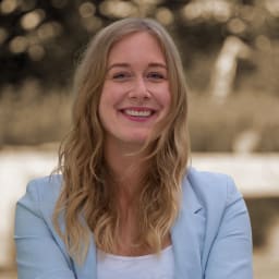 Eine junge Frau mit langen, welligen blonden Haaren, lächelnd warm, draußen mit unscharfen Bäumen im Hintergrund.