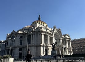 The Palacio de Bellas Artes in Mexico City under a clear blue sky, with a few people walking nearby on a paved area.