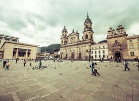Large public square in Bogotá, Colombia, featuring historic colonial buildings and a cathedral under a cloudy sky with people walking and pigeons scattered around.